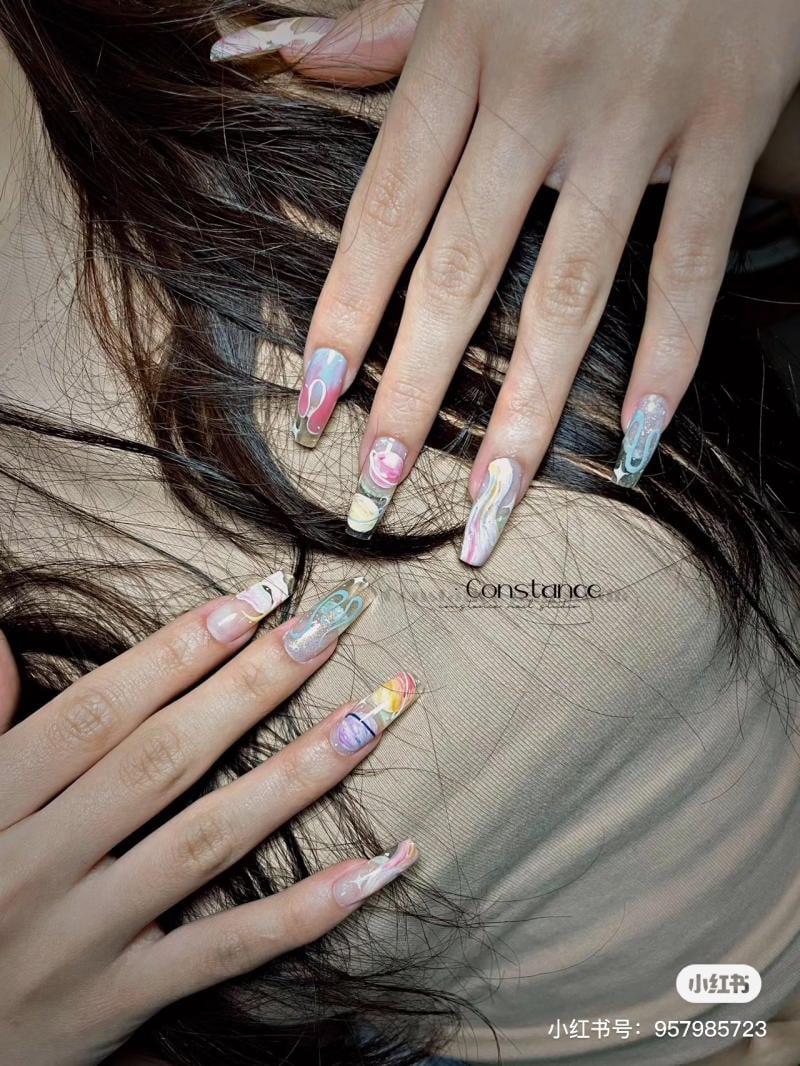 Hai bàn tay với móng tay dài được trang trí nghệ thuật nhiều màu sắc, đặt trên nền tóc và áo màu nâu.