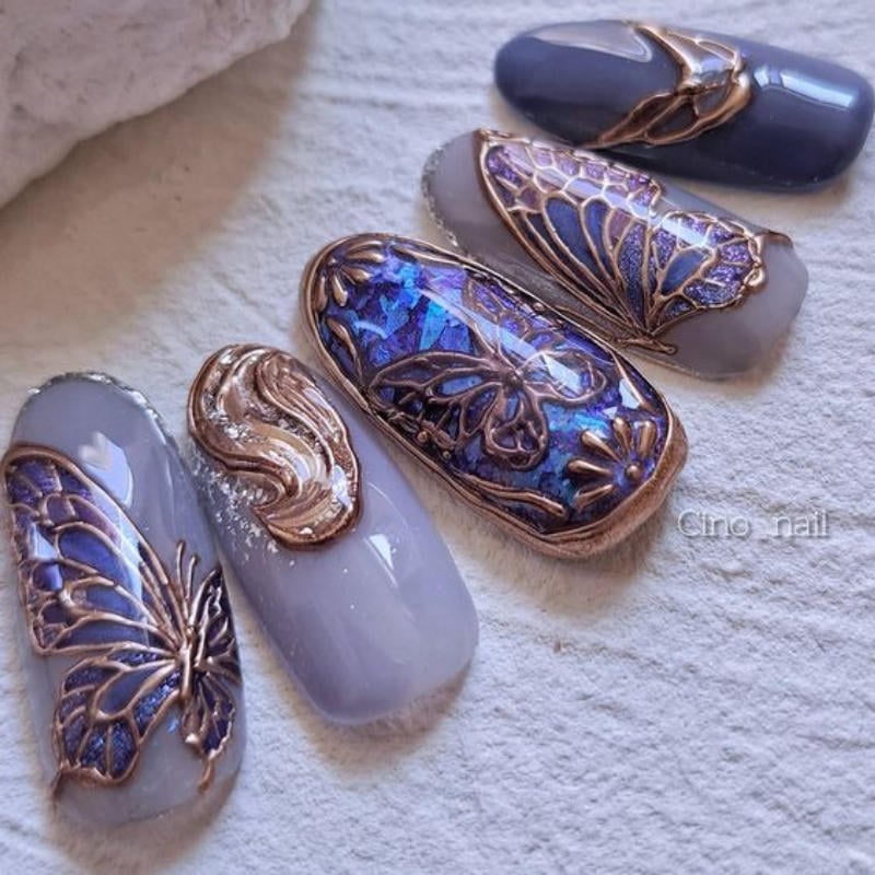 Bộ móng tay giả trang trí hoa văn bướm màu xanh và vàng đồng trên nền màu xám và tím nhạt.