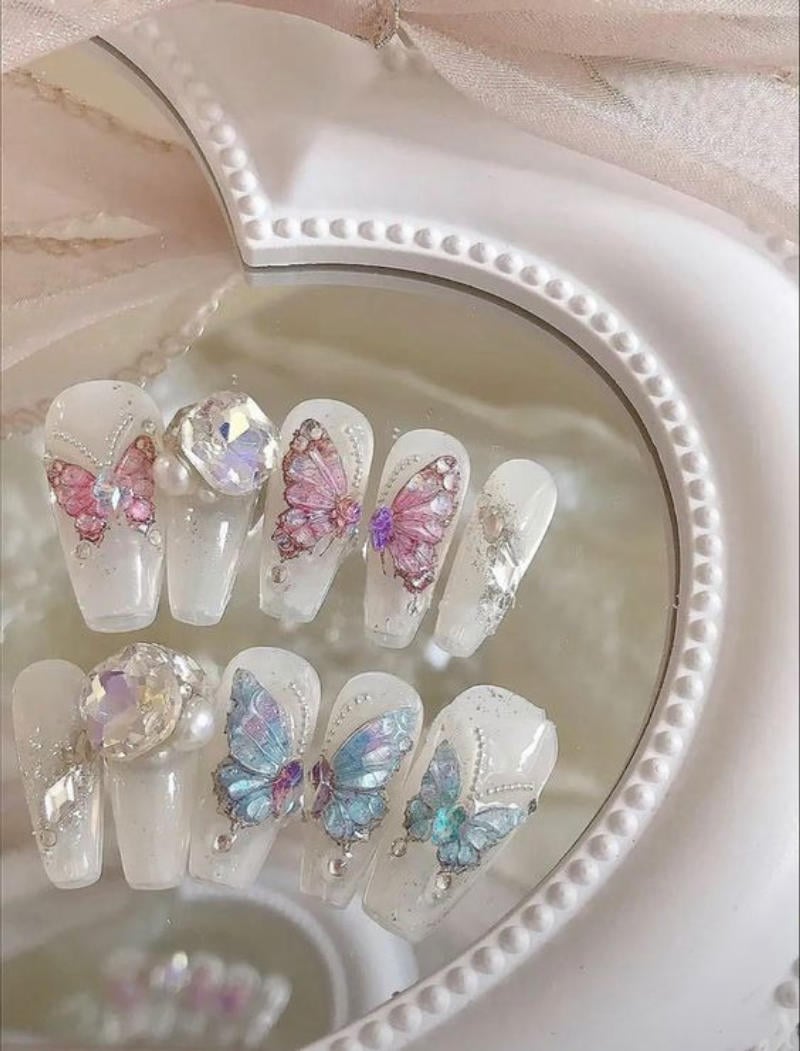 Bộ móng tay giả trang trí với họa tiết bướm màu sắc và đá quý, đặt trên nền gương hình trái tim.