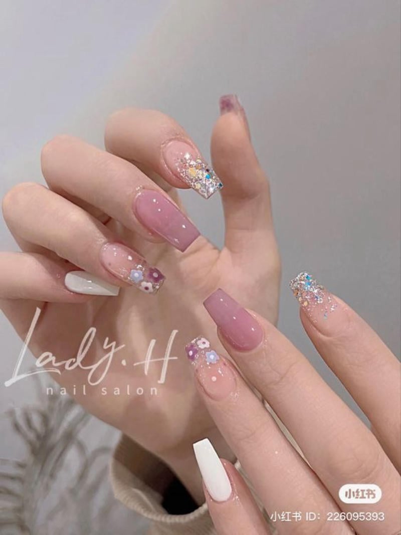 Bàn tay với móng tay được trang trí họa tiết và màu sắc khác nhau, bao gồm màu hồng, trắng và ánh kim tuyến.