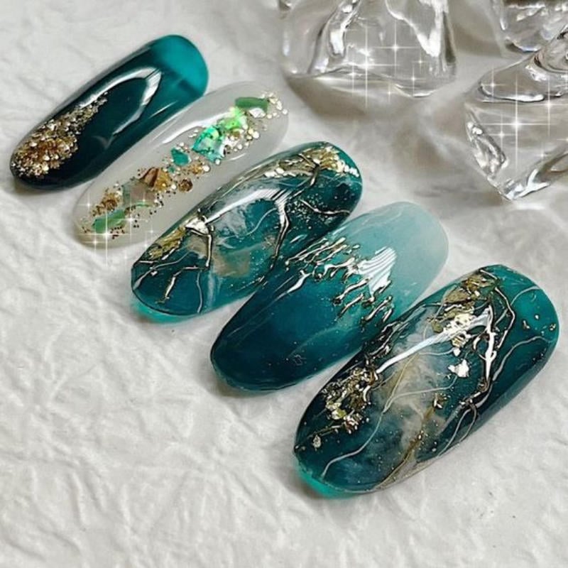 Bộ móng tay giả màu xanh lục, trang trí họa tiết vàng và đá lấp lánh, đặt trên nền trắng.