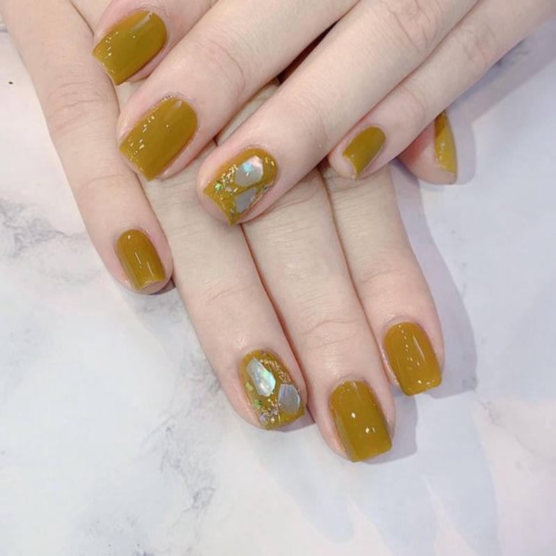 Bàn tay với móng tay sơn màu nâu, hai ngón có đính đá trang trí, đặt trên nền đá cẩm thạch trắng.