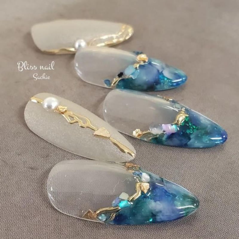 Mẫu móng tay giả trang trí đá xanh, vàng và ngọc trai trên nền trong suốt, đặt trên bề mặt vải xám.