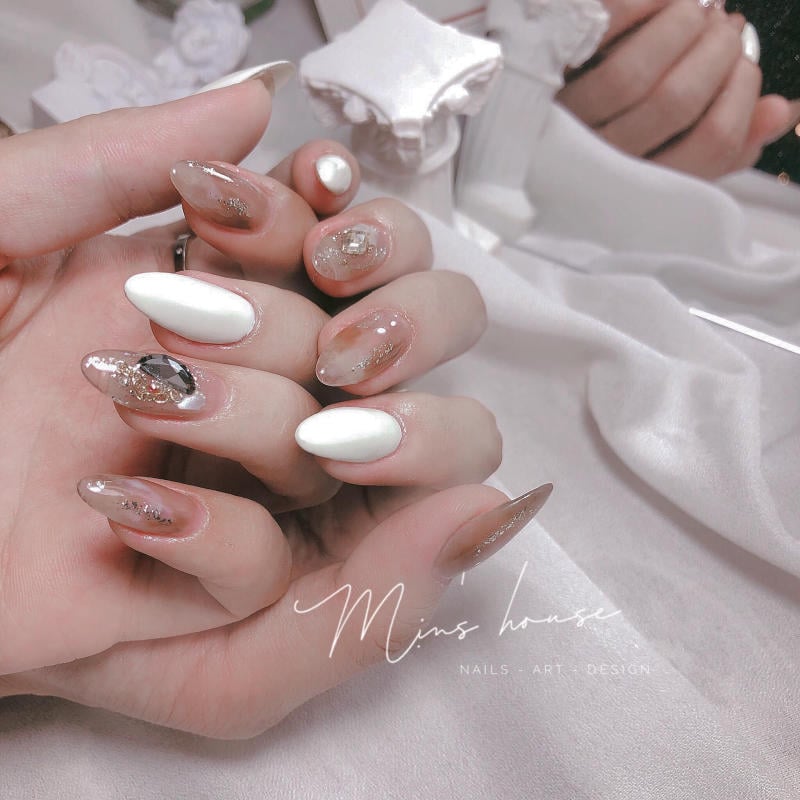 Bàn tay với móng tay được trang trí nghệ thuật, sử dụng màu trắng và các chi tiết lấp lánh trên nền vải trắng.