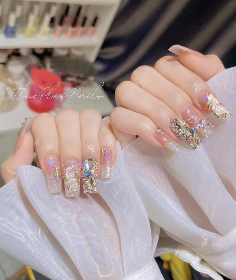 Bàn tay với móng tay dài được trang trí đá quý và họa tiết sáng lấp lánh, nền có kệ sơn móng tay.