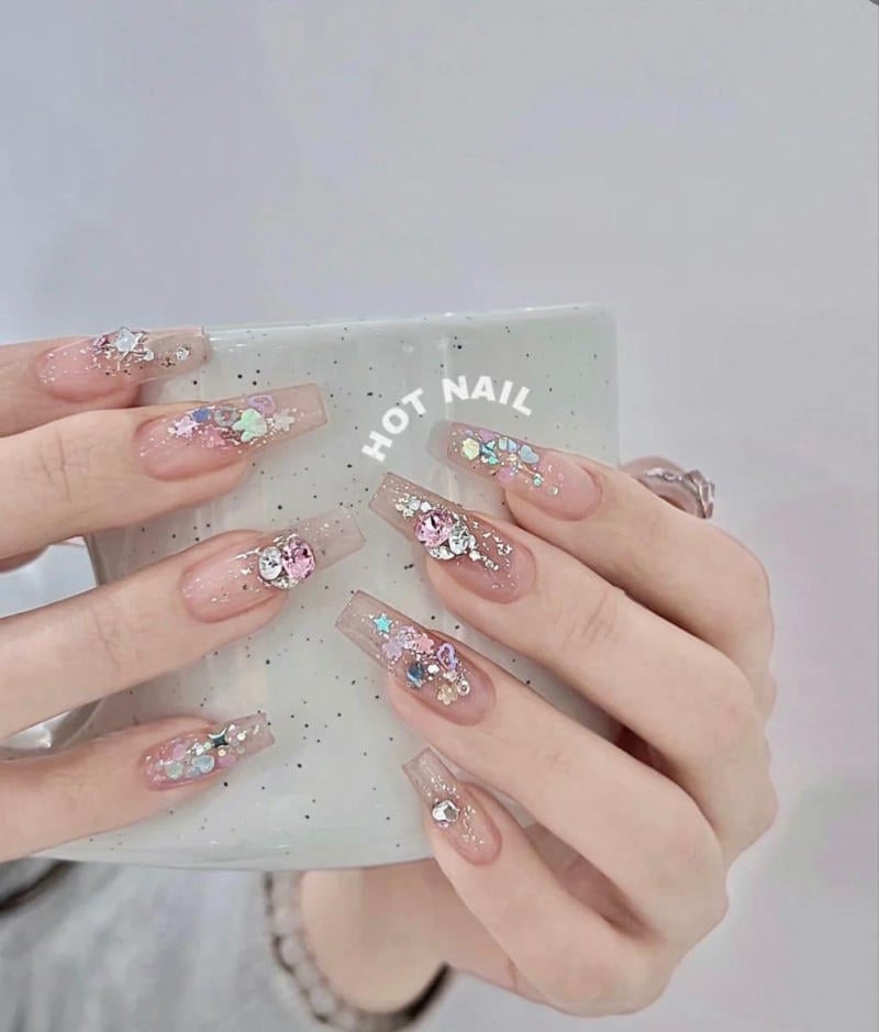 Bàn tay có móng tay dài, trang trí bằng đá lấp lánh, cầm một chiếc cốc màu xám nhạt.