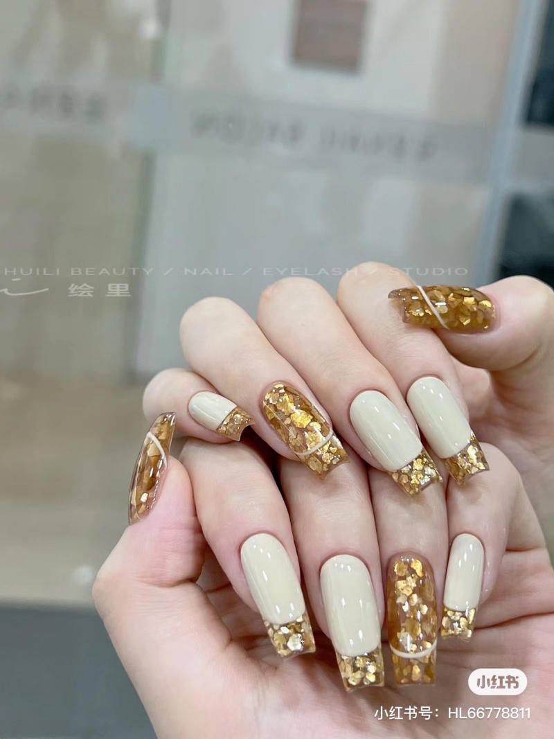 Bàn tay với móng tay dài, sơn màu be và trang trí kim tuyến vàng lấp lánh, nền mờ.