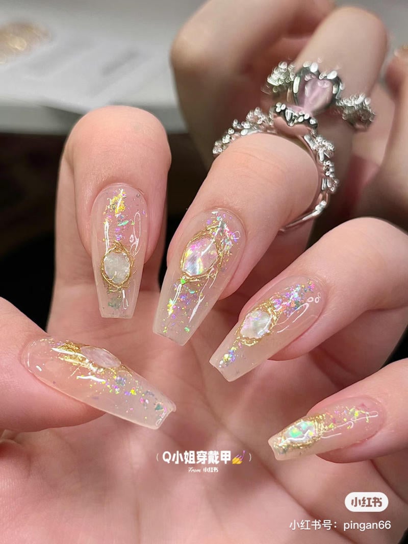 Bàn tay với móng tay dài được trang trí bằng họa tiết vàng và ánh nhũ, đeo nhẫn bạc có đá trang trí.