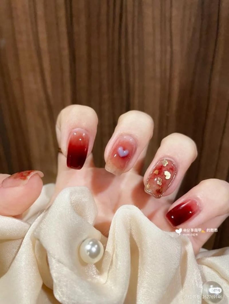 Bàn tay với móng tay trang trí màu đỏ và họa tiết hình trái tim, nền vải màu trắng ngà có nút ngọc trai.