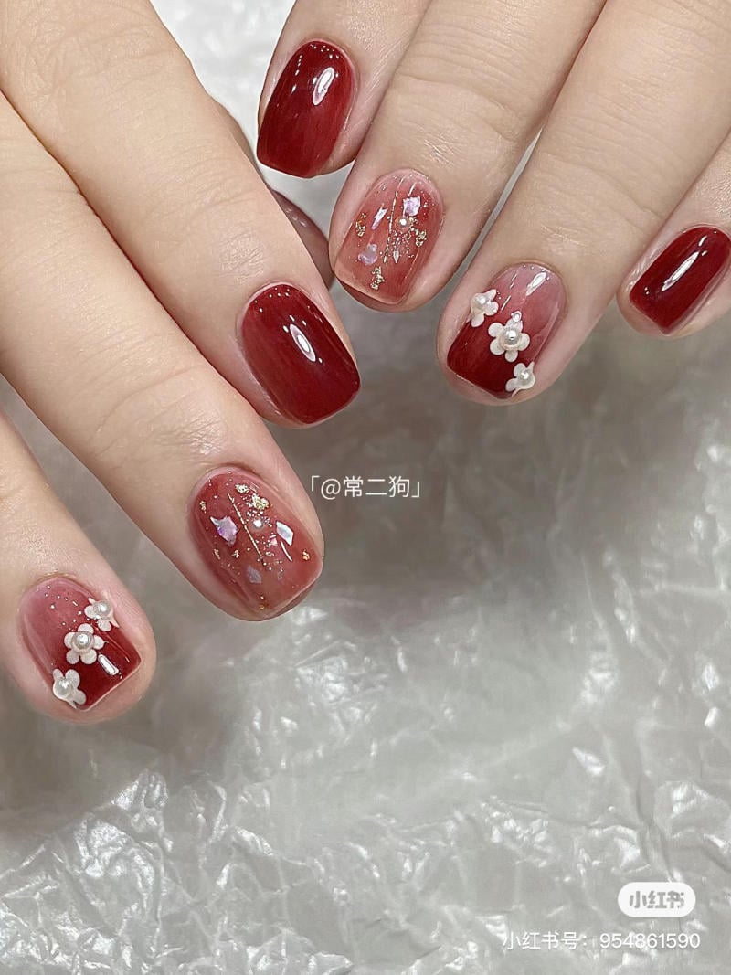 Bàn tay với móng tay sơn đỏ, trang trí hoa trắng và kim tuyến trên nền vải trắng.