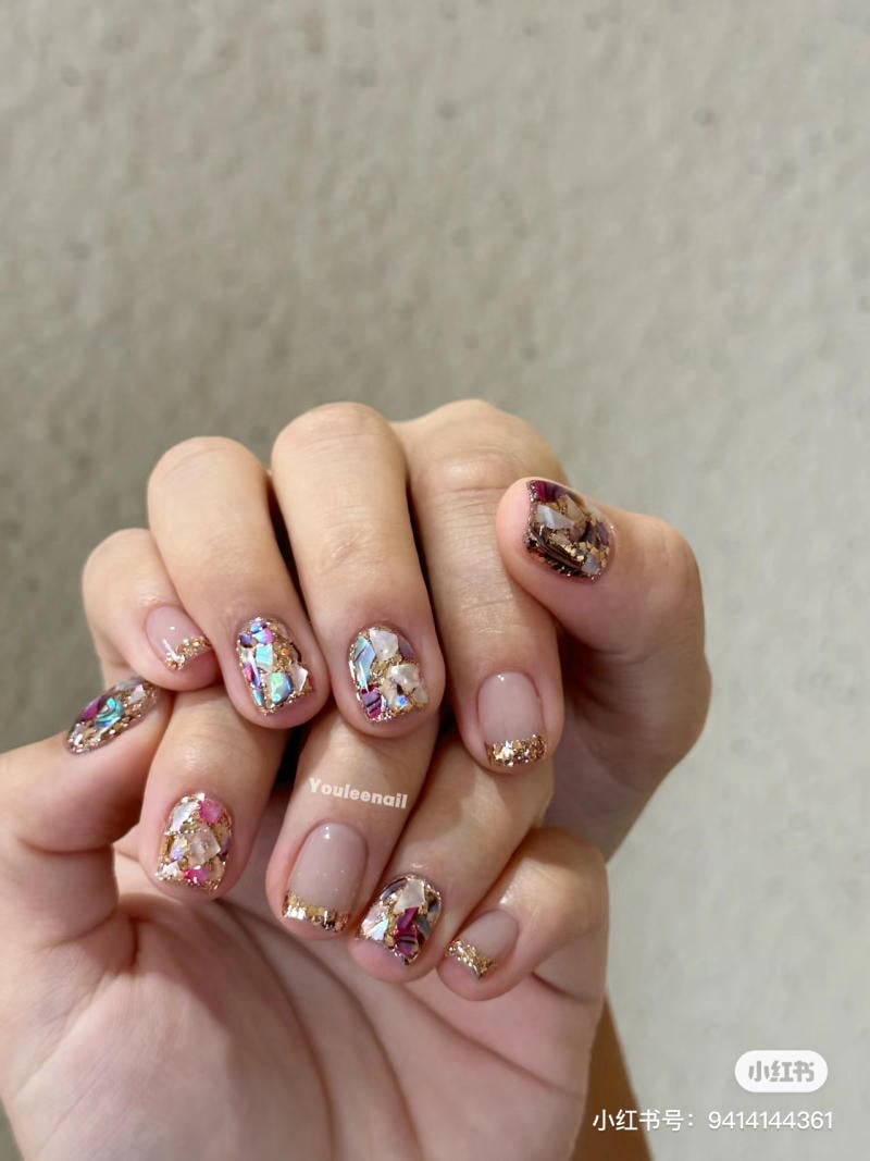 Bàn tay với móng tay được trang trí bằng họa tiết đá màu sắc và viền vàng lấp lánh trên nền móng tự nhiên.