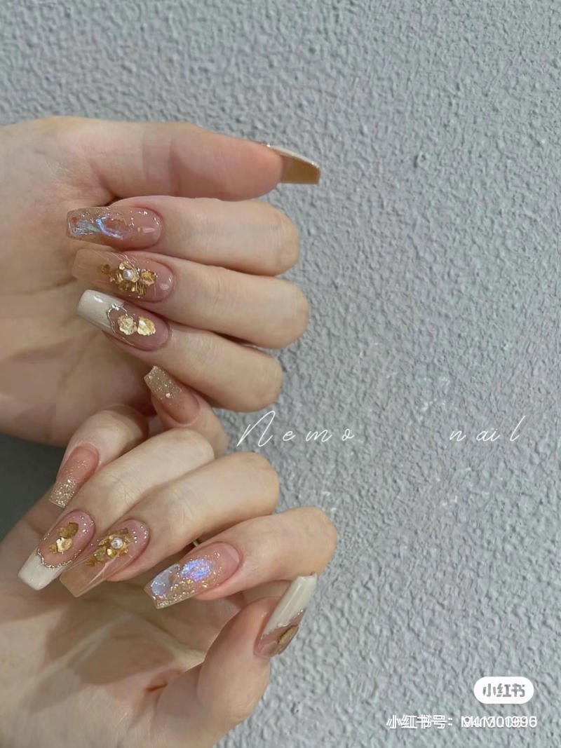 Bàn tay với móng tay được trang trí cầu kỳ bằng màu nude, kim tuyến và họa tiết vàng lấp lánh.