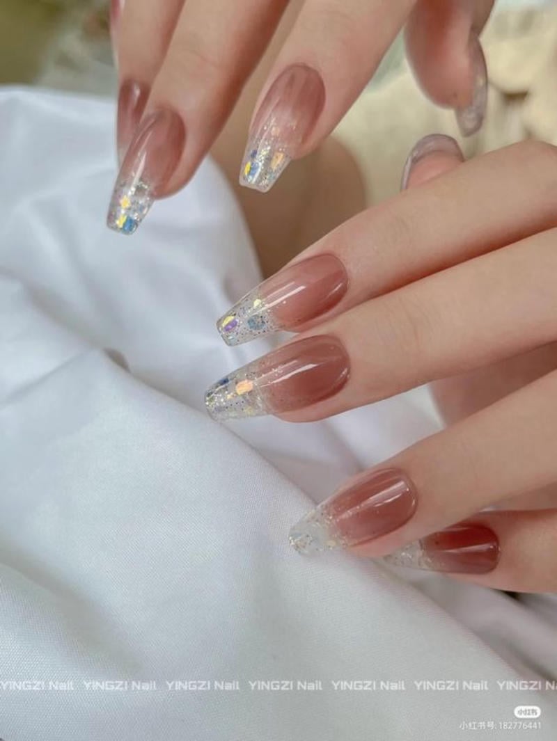 Bàn tay với móng tay dài, sơn màu nâu nhạt kết hợp kim tuyến lấp lánh, đặt trên nền vải trắng.