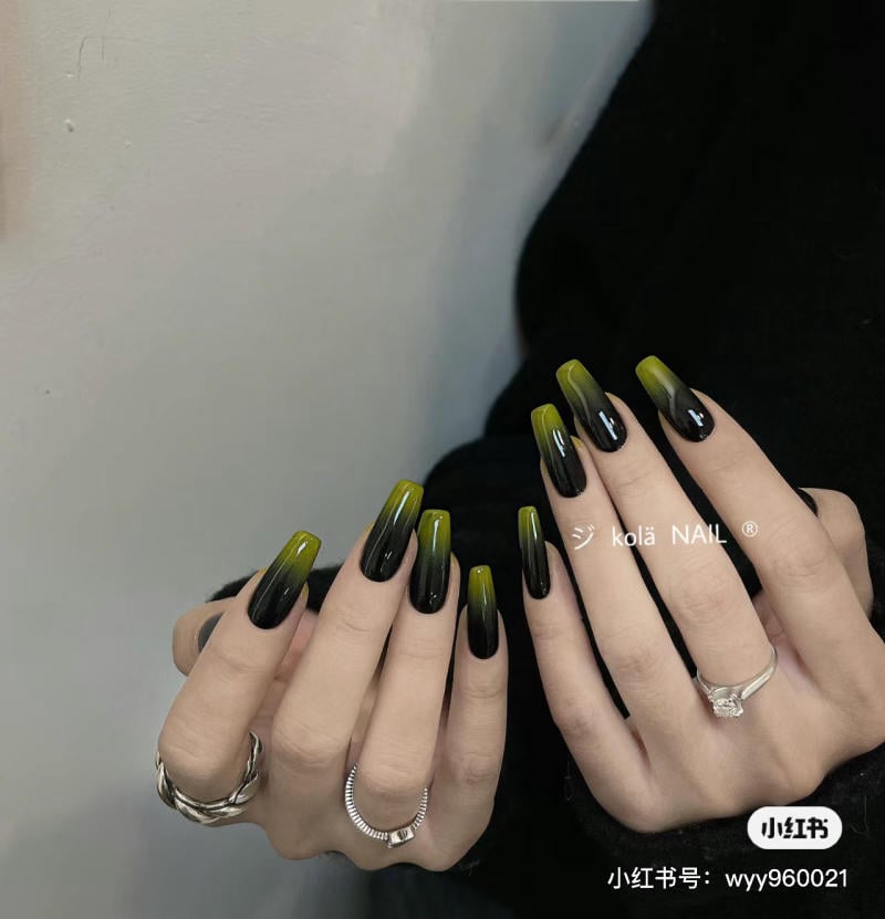 Bàn tay với móng tay dài màu đen và xanh lá, đeo nhẫn bạc, trên nền tường trắng và trang phục tối màu.