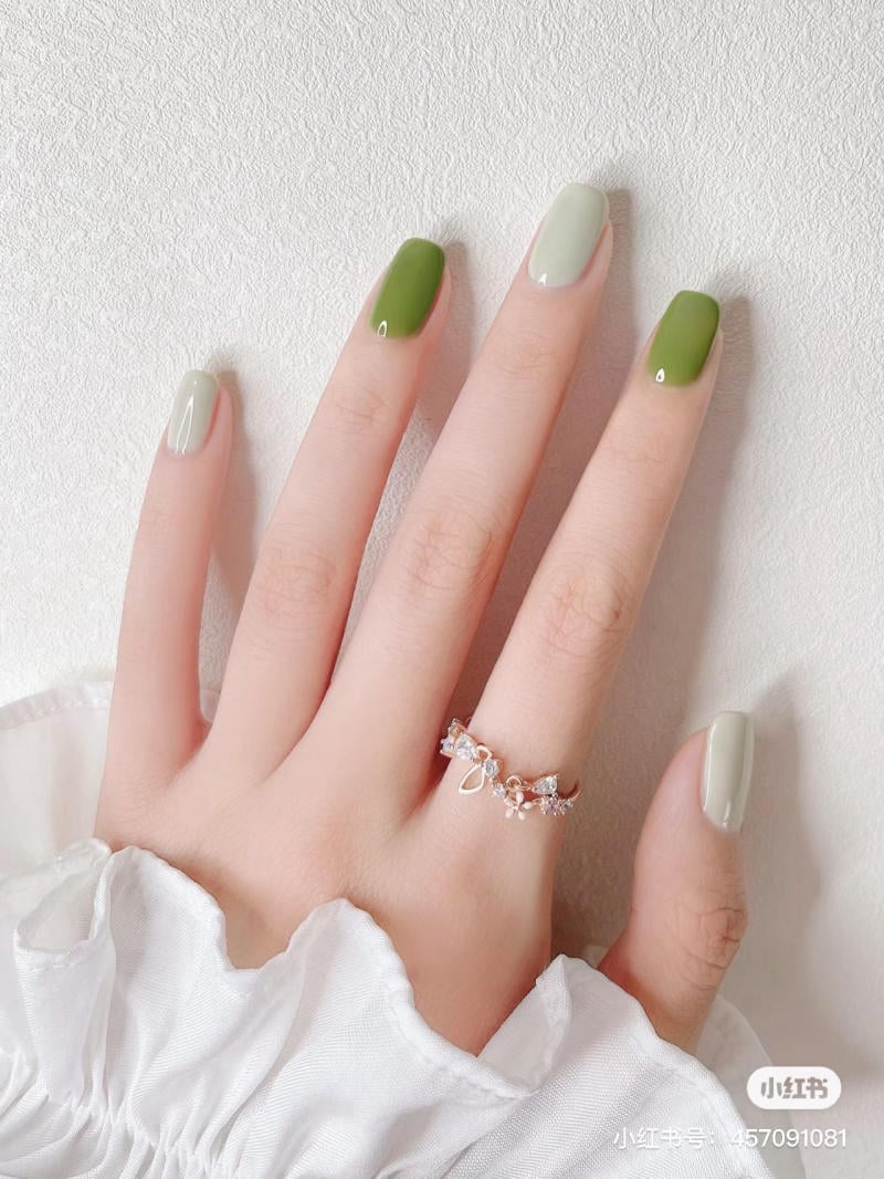 Bàn tay với móng tay sơn màu xanh lá cây và xám, đeo nhẫn trang trí, trên nền vải trắng.
