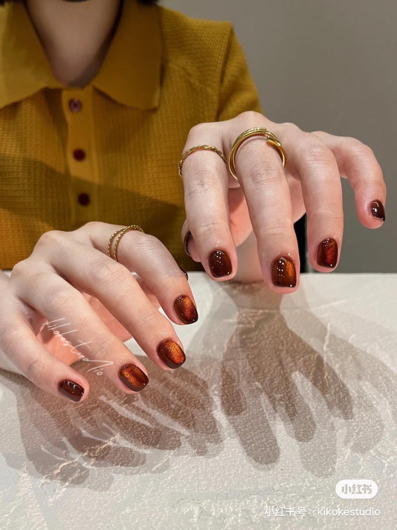 Đ&ocirc;i tay với m&oacute;ng sơn m&agrave;u n&acirc;u &oacute;ng &aacute;nh, đeo nhiều nhẫn v&agrave;ng, đặt tr&ecirc;n bề mặt c&oacute; &aacute;nh s&aacute;ng phản chiếu.