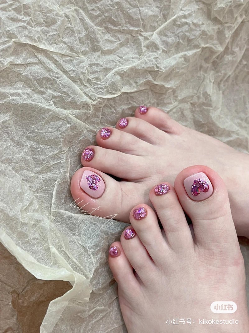 B&agrave;n ch&acirc;n với m&oacute;ng được sơn m&agrave;u hồng v&agrave; trang tr&iacute; kim tuyến, đặt tr&ecirc;n nền giấy nhăn m&agrave;u s&aacute;ng.