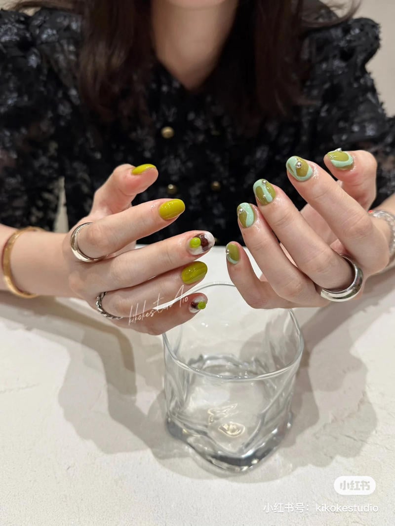 Người đang khoe bộ m&oacute;ng tay sơn m&agrave;u xanh l&aacute; v&agrave; xanh nhạt, cầm một ly thủy tinh tr&ecirc;n b&agrave;n.