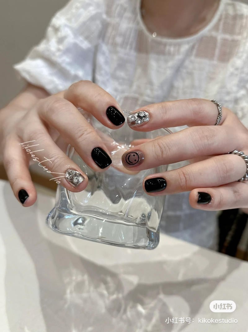 Hai b&agrave;n tay với m&oacute;ng tay đen v&agrave; họa tiết, cầm một chiếc ly thủy tinh trong suốt, nền vải trắng.