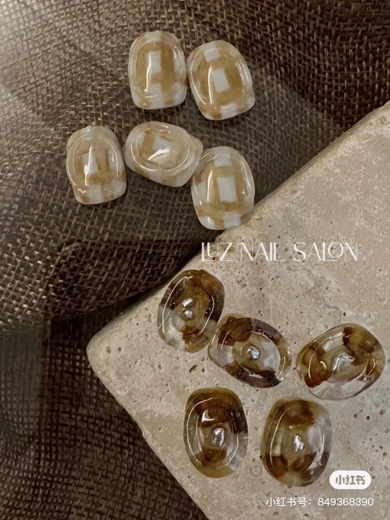 Bộ m&oacute;ng giả m&agrave;u n&acirc;u v&agrave; trắng, đặt tr&ecirc;n nền vải v&agrave; đ&aacute;, với d&ograve;ng chữ "Luz Nail Salon".