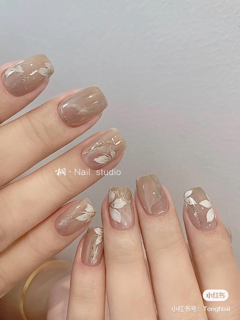 B&agrave;n tay với m&oacute;ng tay được sơn m&agrave;u nude, trang tr&iacute; hoa trắng v&agrave; nhũ v&agrave;ng. M&oacute;ng c&oacute; h&igrave;nh vu&ocirc;ng v&agrave; d&agrave;i vừa phải.