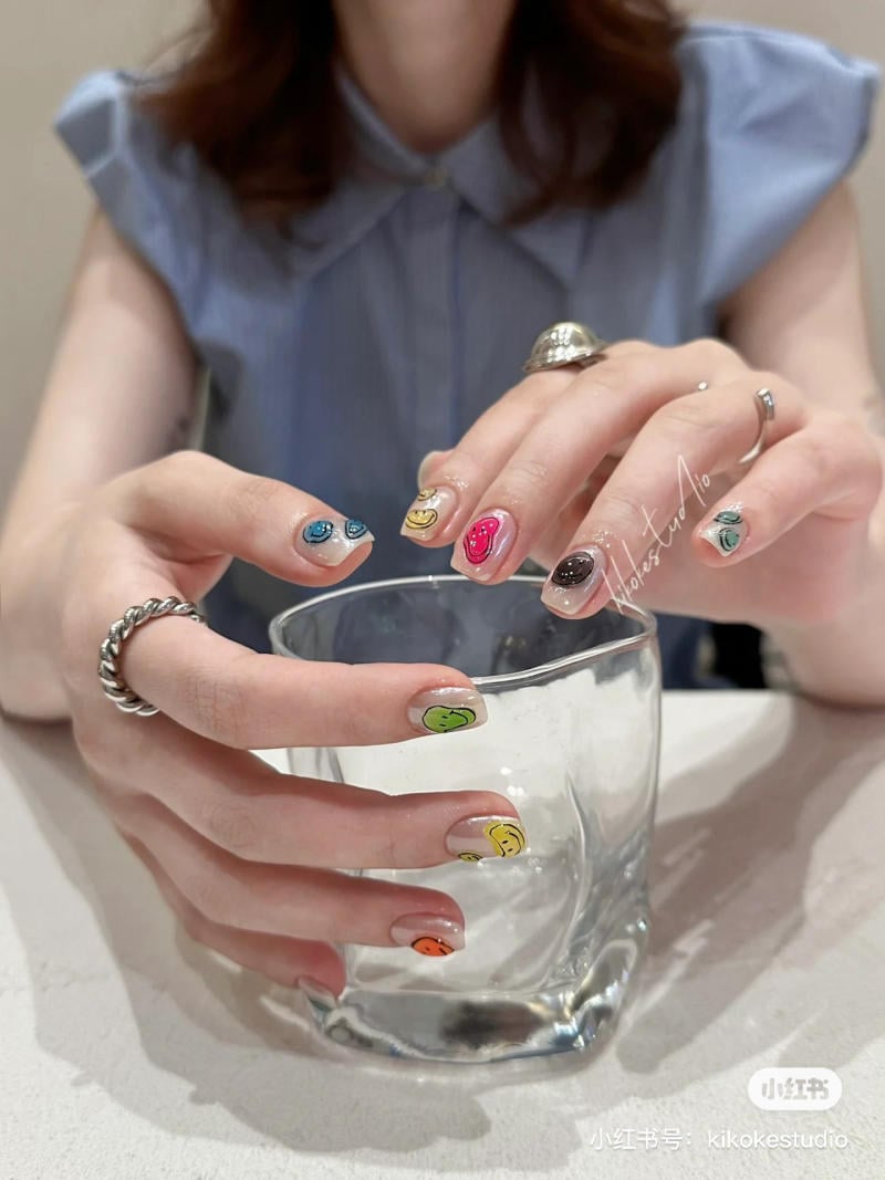 Người với m&oacute;ng tay vẽ họa tiết mặt cười đa sắc, cầm ly thủy tinh tr&ecirc;n b&agrave;n trắng.