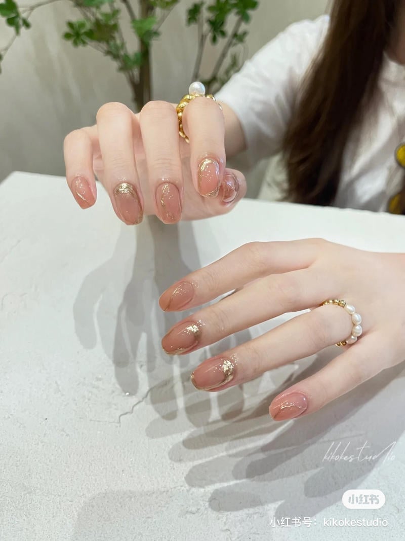 B&agrave;n tay với m&oacute;ng tay nghệ thuật m&agrave;u n&acirc;u nhạt, trang tr&iacute; nhũ v&agrave;ng, đeo nhẫn ngọc trai v&agrave; v&agrave;ng.