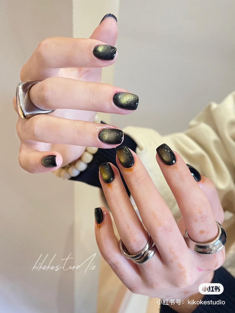 B&agrave;n tay với m&oacute;ng tay được sơn m&agrave;u đen v&agrave; xanh l&aacute; lấp l&aacute;nh, đeo nhiều nhẫn bạc, tạo d&aacute;ng tr&ecirc;n nền m&agrave;u nhạt.