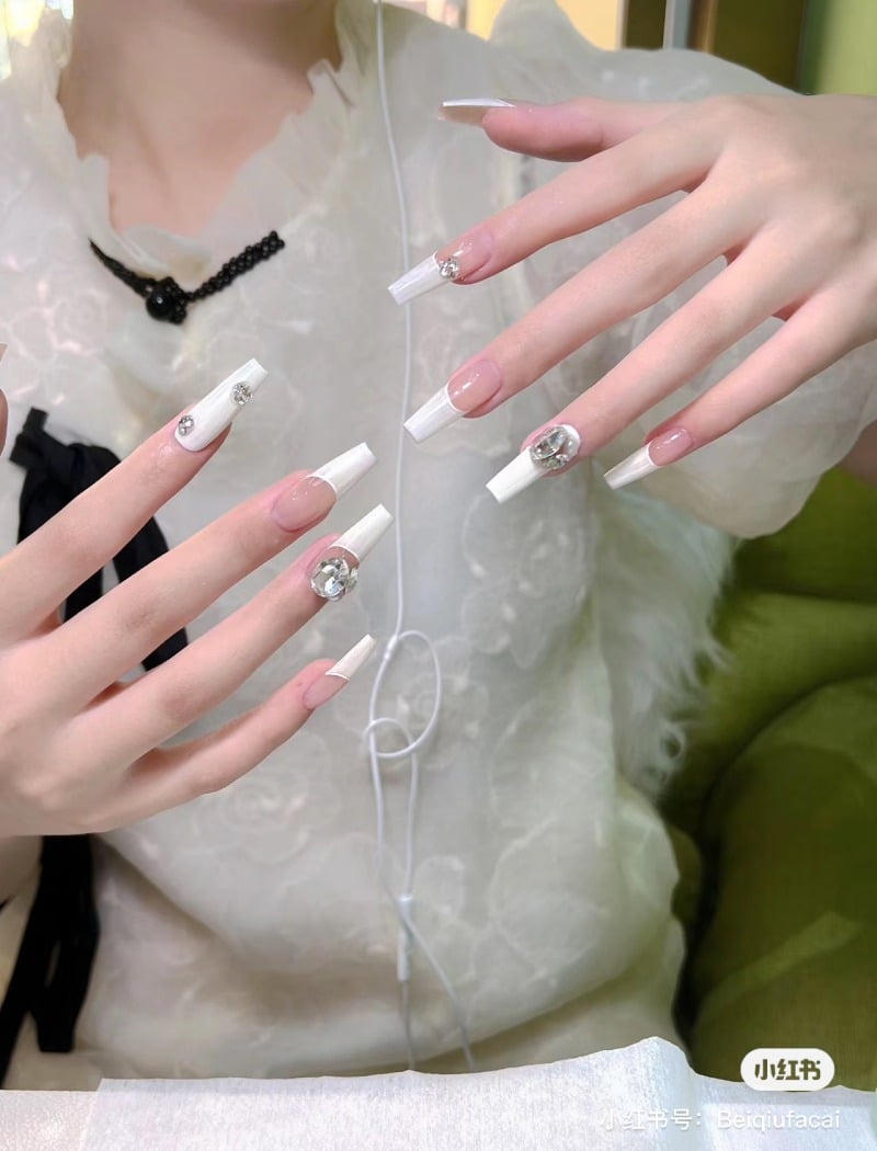 Bàn tay với móng tay dài, trang trí đá lấp lánh, trên nền áo trắng có hoa văn tinh tế.