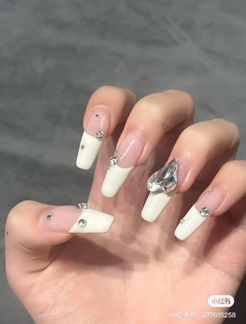 Bàn tay với móng tay dài được sơn màu trắng và trang trí bằng đá lấp lánh, trên nền xám.