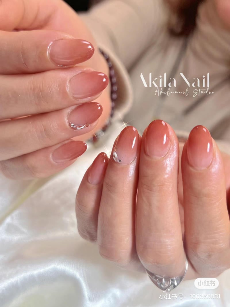 Bàn tay với móng tay sơn màu nâu nhạt, có đính đá trang trí, nền vải trắng.