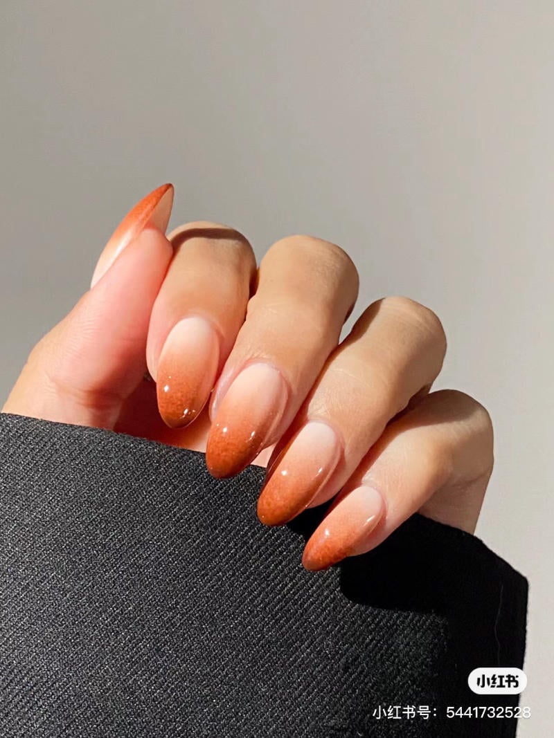 Bàn tay với móng tay sơn màu cam ombre, cầm một mảnh vải màu đen, nền xám nhạt.