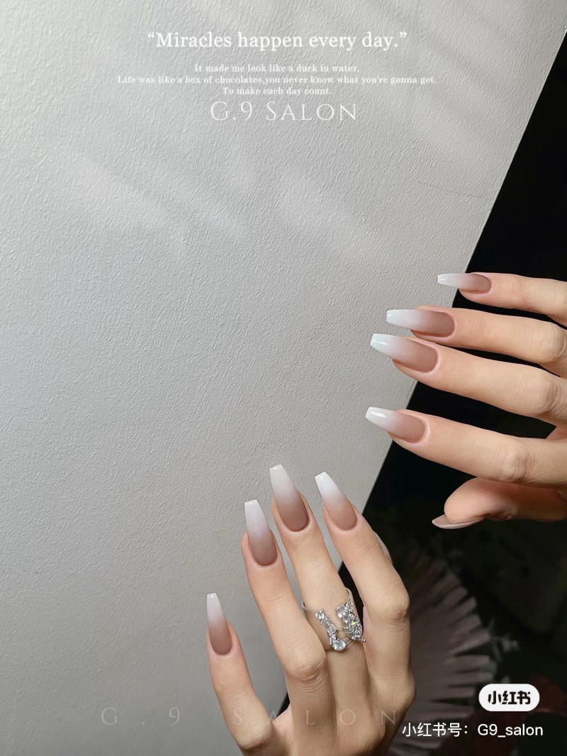 Bàn tay với móng tay dài sơn ombre trắng, đeo nhẫn, trên nền tường trắng có chữ.