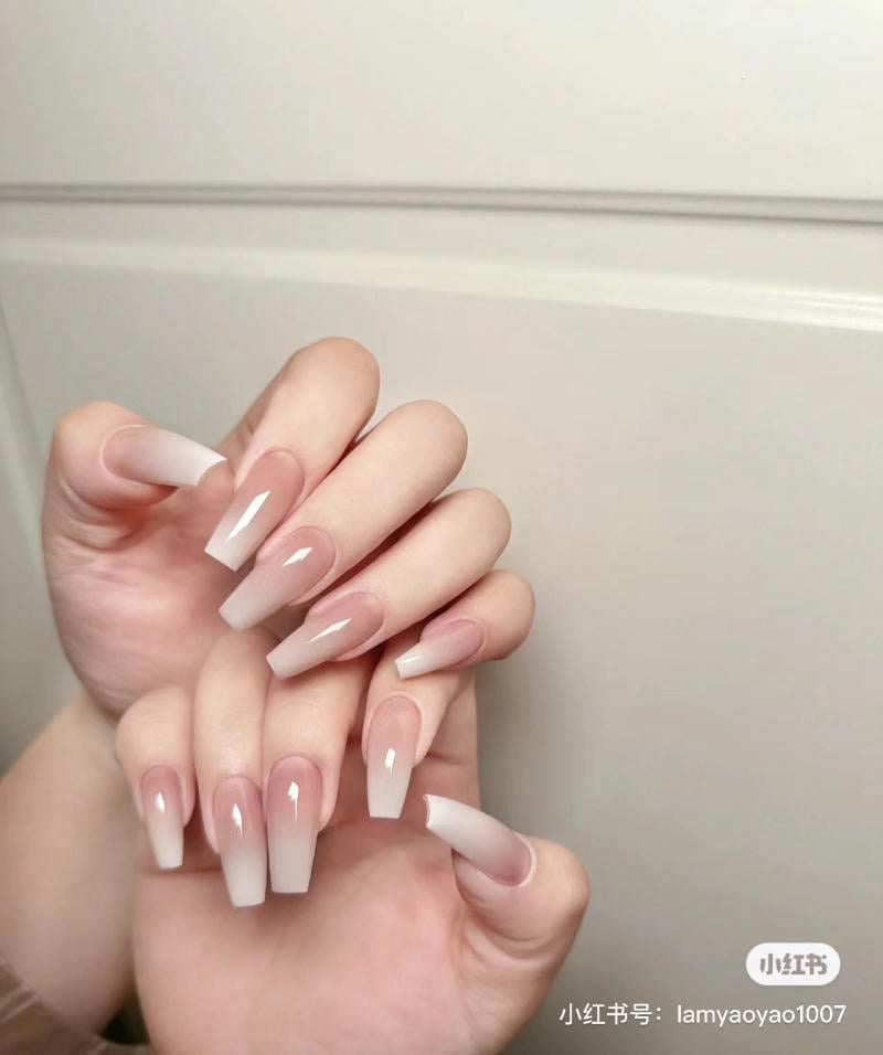 Bàn tay với móng tay dài, được sơn màu hồng nhạt, tạo kiểu ombre trắng, nền màu trắng nhạt.