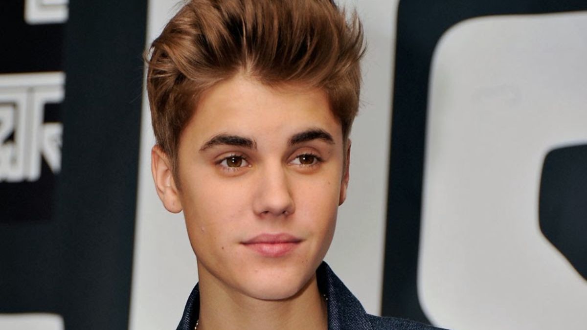 toc-justin-bieber-19 Người trẻ tóc nâu, mặc áo khoác tối màu, đứng trước phông nền có họa tiết chữ.