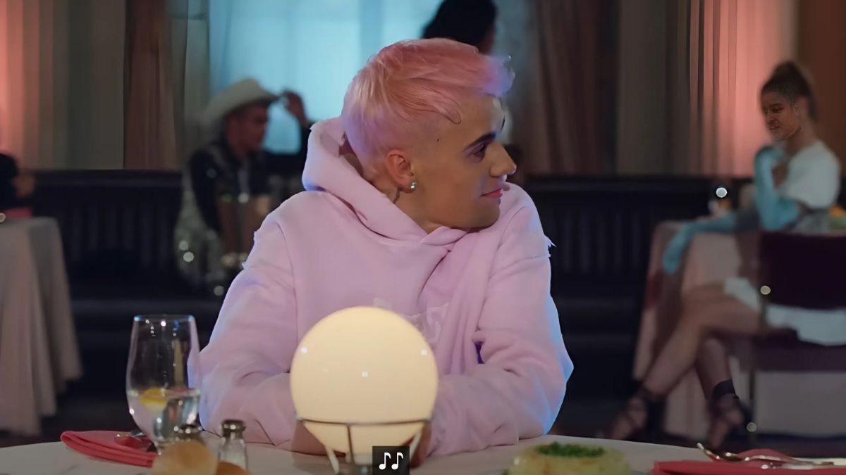 toc-justin-bieber-23 Người với tóc hồng ngồi trong nhà hàng, mặc áo hoodie hồng, trước mặt có đèn và ly nước.