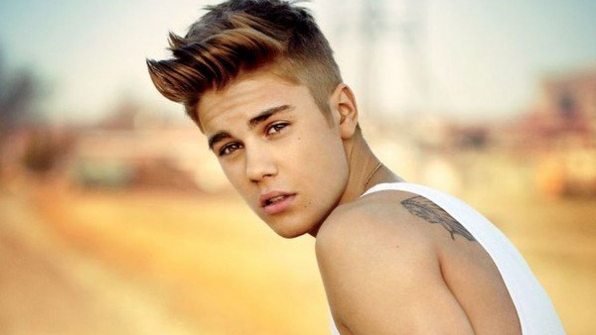 toc-justin-bieber-27 Người đàn ông trẻ mặc áo trắng, tóc ngắn, nhìn về phía máy ảnh, bối cảnh mờ nhạt phía sau.