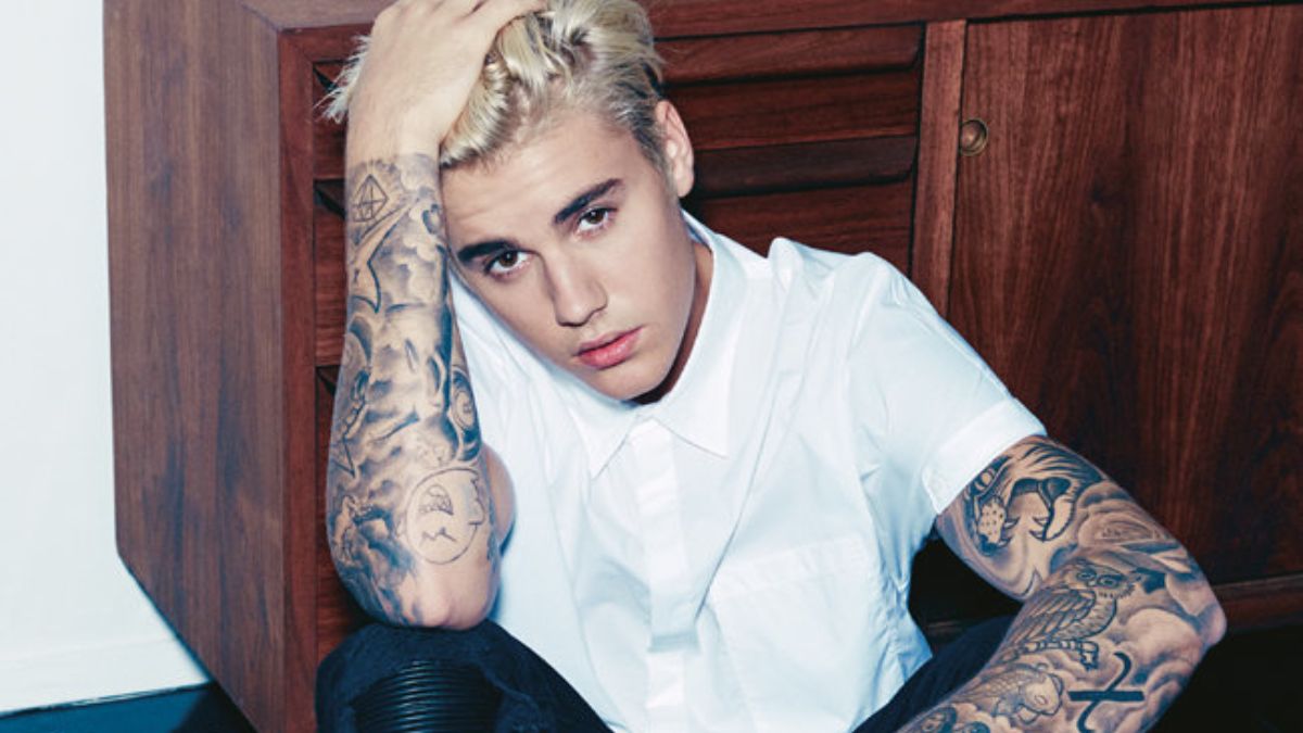 toc-justin-bieber-28 Người đàn ông tóc vàng, mặc áo sơ mi trắng, ngồi tựa vào tủ gỗ, tay có nhiều hình xăm nghệ thuật.