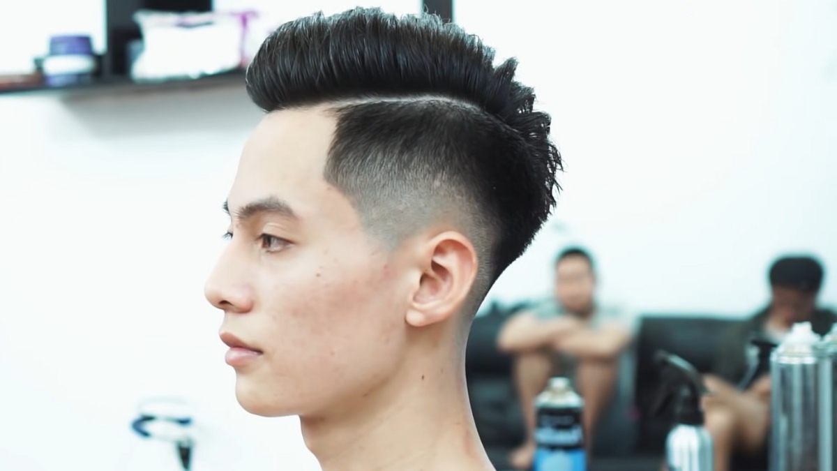 Người có kiểu tóc hiện đại với hai bên cạo ngắn, đứng trong một tiệm cắt tóc với người khác ở phía xa.