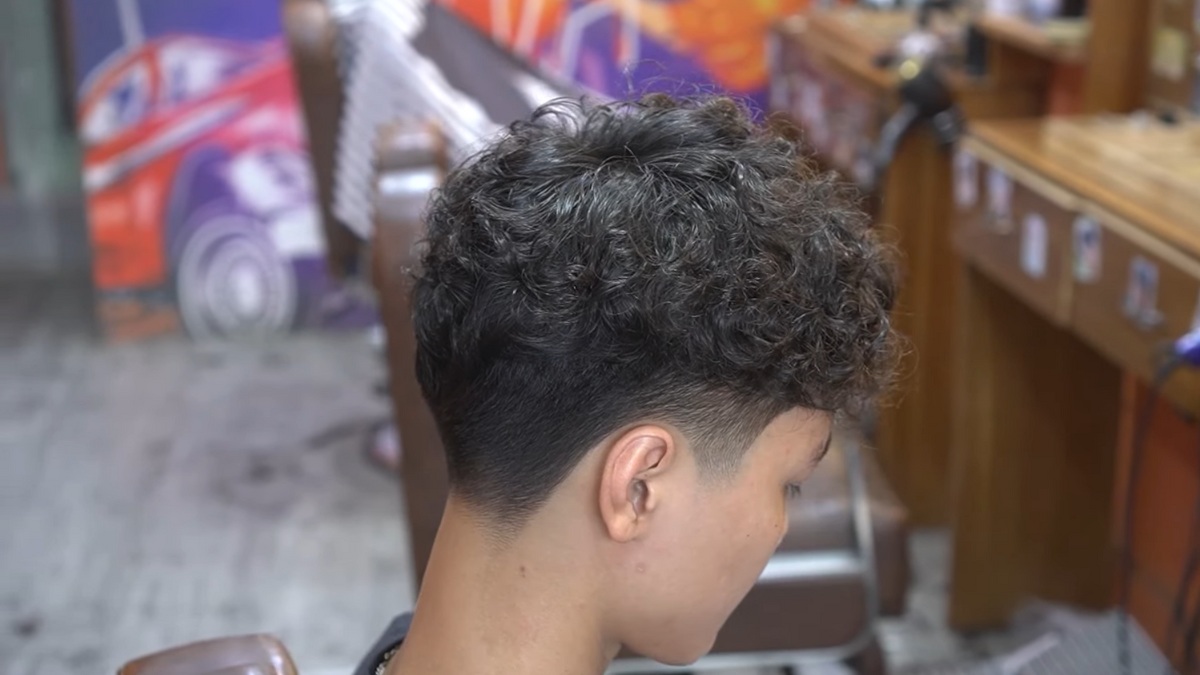 Người đang ngồi trong tiệm cắt tóc, kiểu tóc xoăn ngắn, cắt sát hai bên, không gian tiệm có trang trí màu sắc.