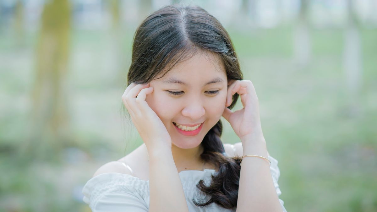 Người phụ nữ trẻ cười tươi, tay chạm vào tóc, đứng ngoài trời với nền cây xanh.