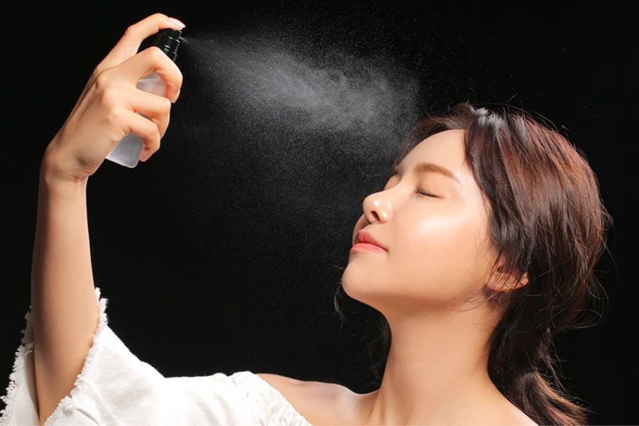 Người phụ nữ đang xịt khoáng lên mặt từ chai xịt nhỏ, nền đen phía sau.