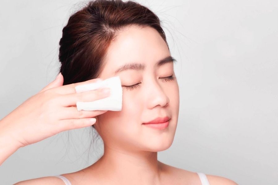 Người phụ nữ đang d&ugrave;ng b&ocirc;ng tẩy trang lau mặt, mắt nhắm thư gi&atilde;n, nền trắng.