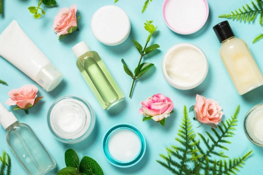 3-cac-loai-my-pham-xanh-pho-bien_33cb63d343e04abc886542d9485f4587 Các lọ mỹ phẩm và hoa hồng trên nền xanh, sắp xếp xen kẽ với lá cây xanh tươi.