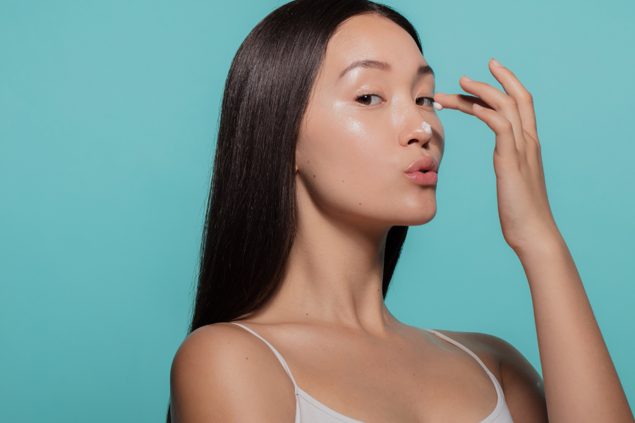 Người phụ nữ chạm tay vào mũi với một vệt kem, nền xanh lam, tóc dài và mặc áo dây.