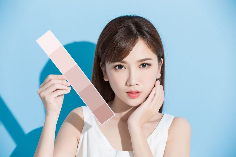 Người phụ nữ cầm bảng màu da, đứng trước nền xanh, tay trái chạm nhẹ lên má, ánh sáng chiếu sáng khuôn mặt.