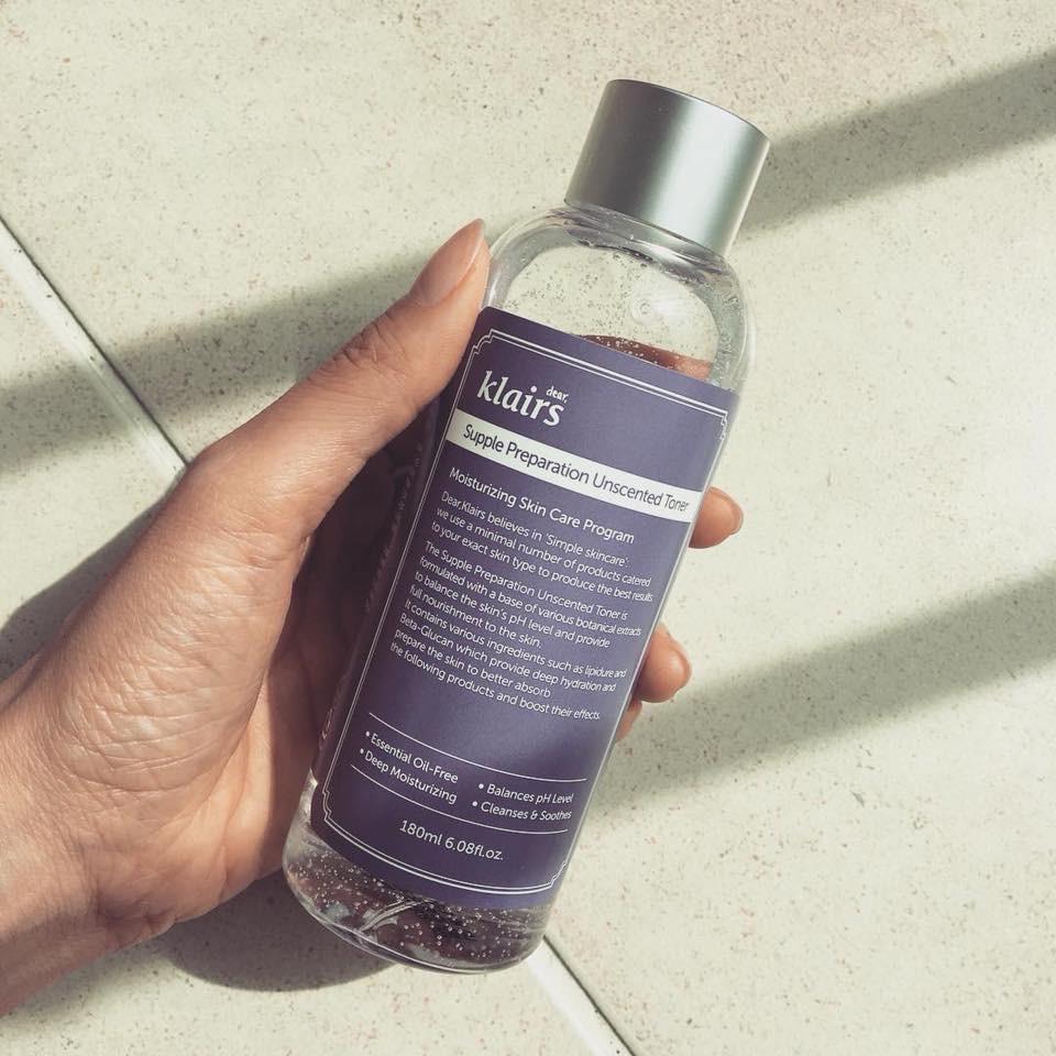 Người cầm chai toner c&oacute; nắp m&agrave;u bạc, nh&atilde;n m&agrave;u t&iacute;m, tr&ecirc;n nền gạch s&aacute;ng m&agrave;u.