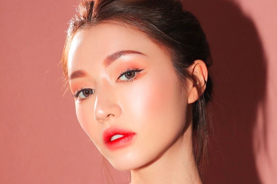 Người mẫu trang điểm nhẹ nh&agrave;ng với t&ocirc;ng m&agrave;u cam hồng, nền hồng nhạt, &aacute;nh s&aacute;ng chiếu từ b&ecirc;n phải.