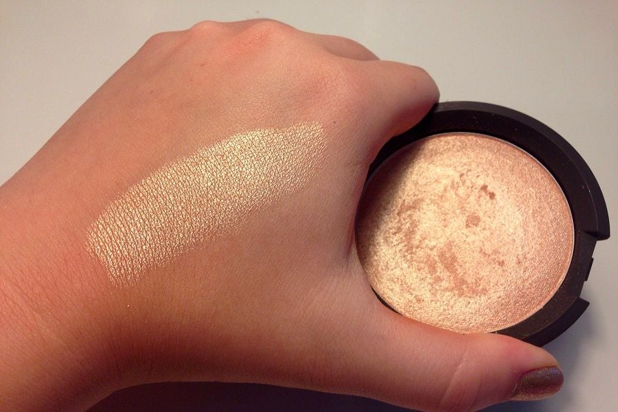 B&agrave;n tay với vệt phấn highlight m&agrave;u v&agrave;ng đồng tr&ecirc;n da, b&ecirc;n cạnh hộp phấn tr&ograve;n c&ugrave;ng m&agrave;u.