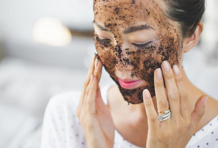 Người phụ nữ thoa mặt nạ dưỡng da tự nhiên màu nâu lên mặt, tay chạm nhẹ vào má, mặc áo trắng.